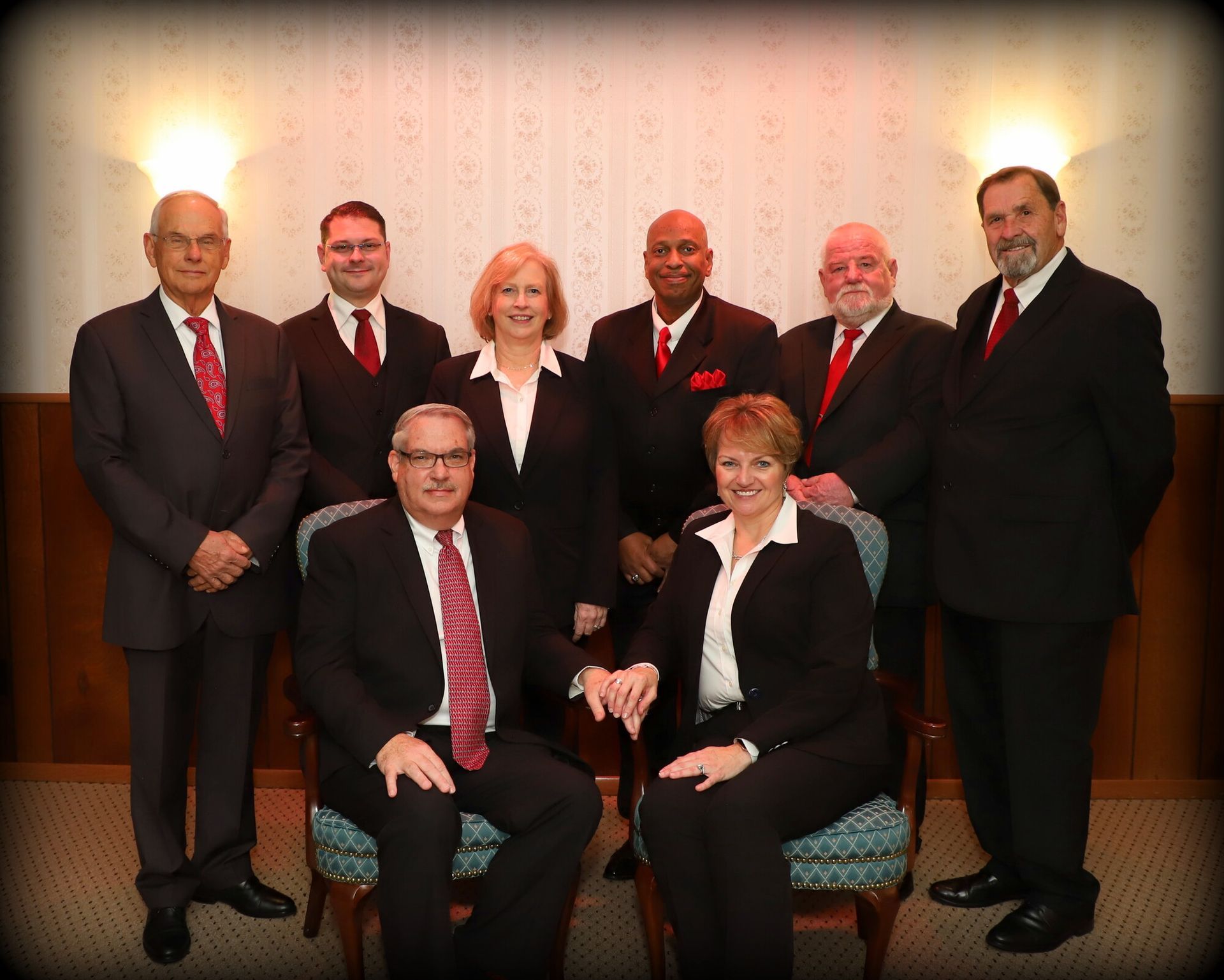 Seven people in business attire posed indoors, with two seated in front and five standing behind them.