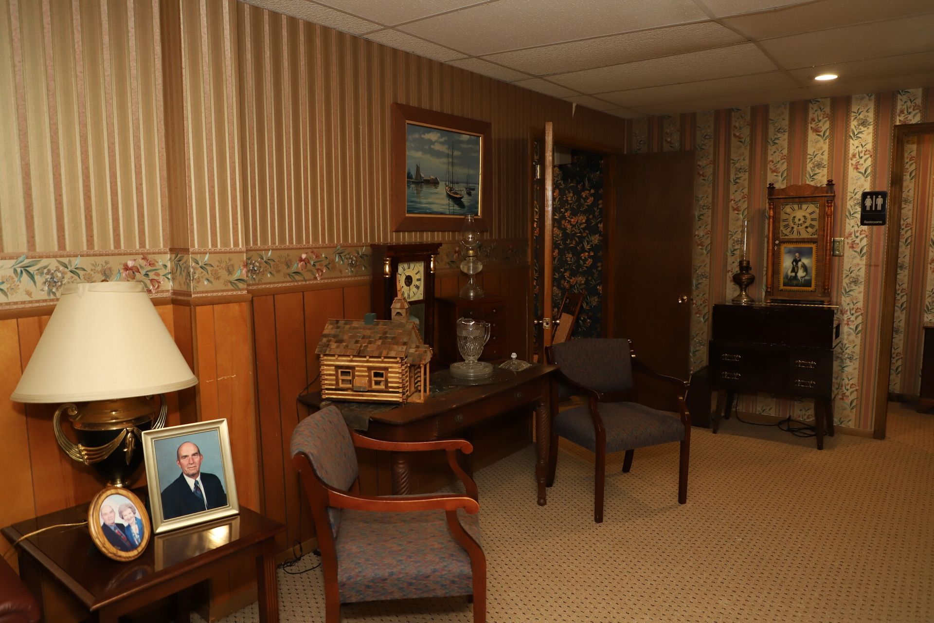 Dim vintage room with wood paneling, patterned wallpaper, chairs, table, lamp, and fireplace.