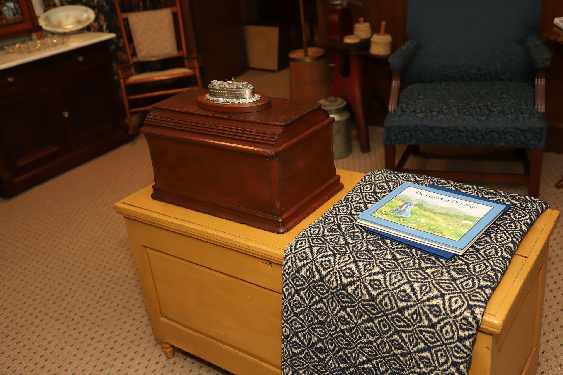 A room with a wooden chest, patterned table runner, and framed artwork on top of a beige cabinet.