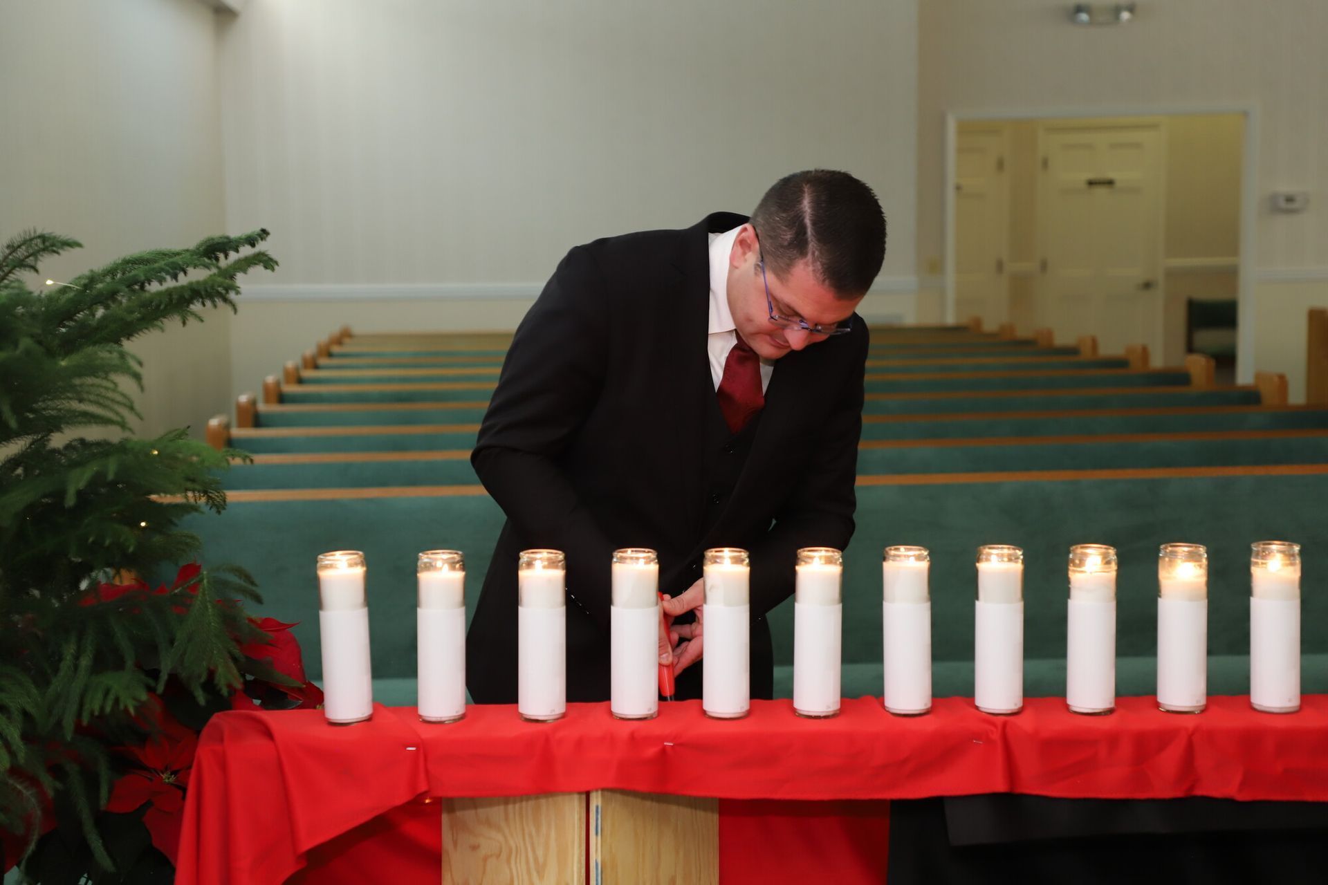 Man in black suit lighting white memorial candles on a red-draped table in a church sanctuary