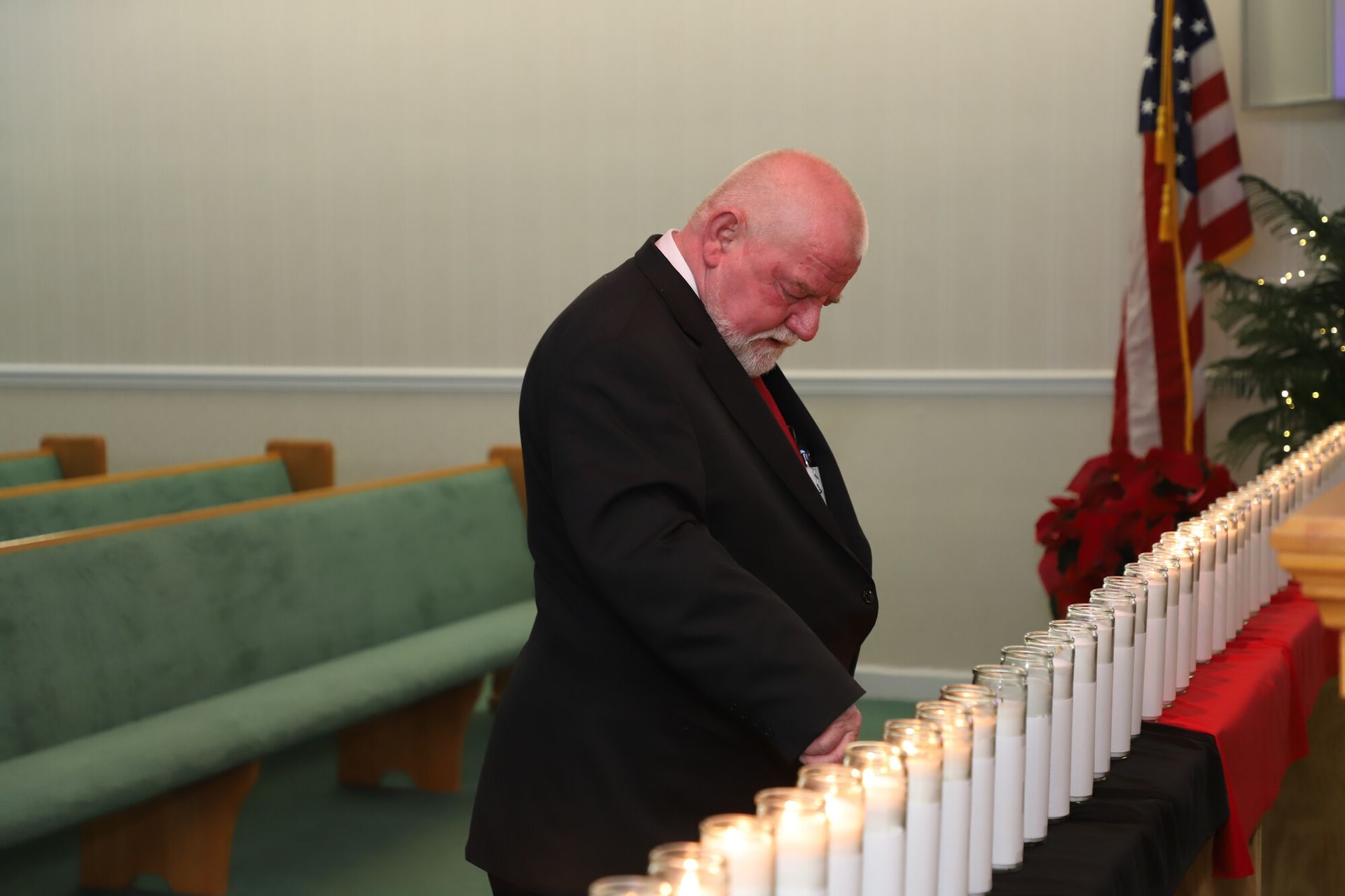 Man in black suit lighting candles in a church beside a red cloth and American flag