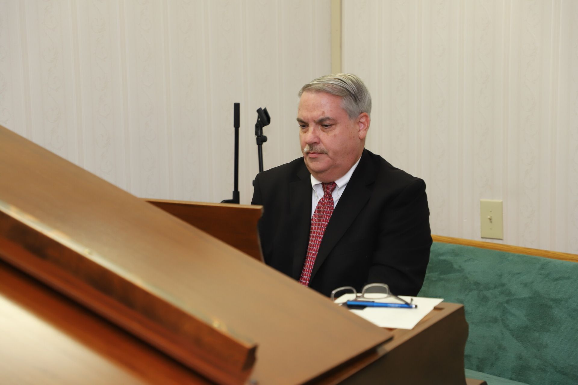 Man in a suit seated at a desk in a formal meeting room, with a green upholstered chair behind him.