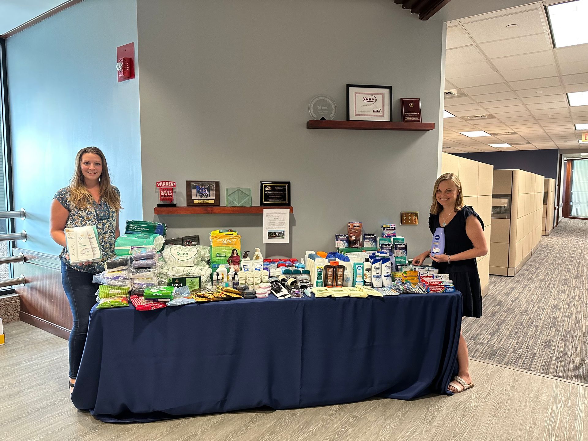 Kristi and Chelsea stand on either side of a table piled with personal items in the MBK lobby.
