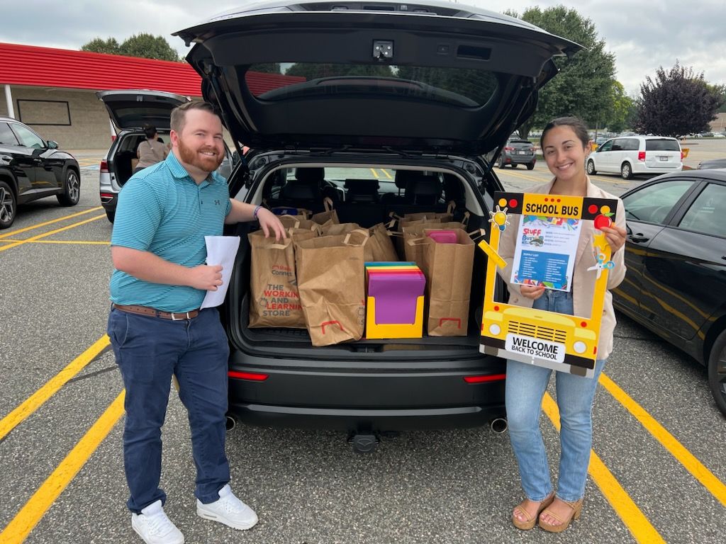 Chis and Olivia stand on either side of an open hatchback, the trunk filled with bags of schools supplies to be delivered to United Way.