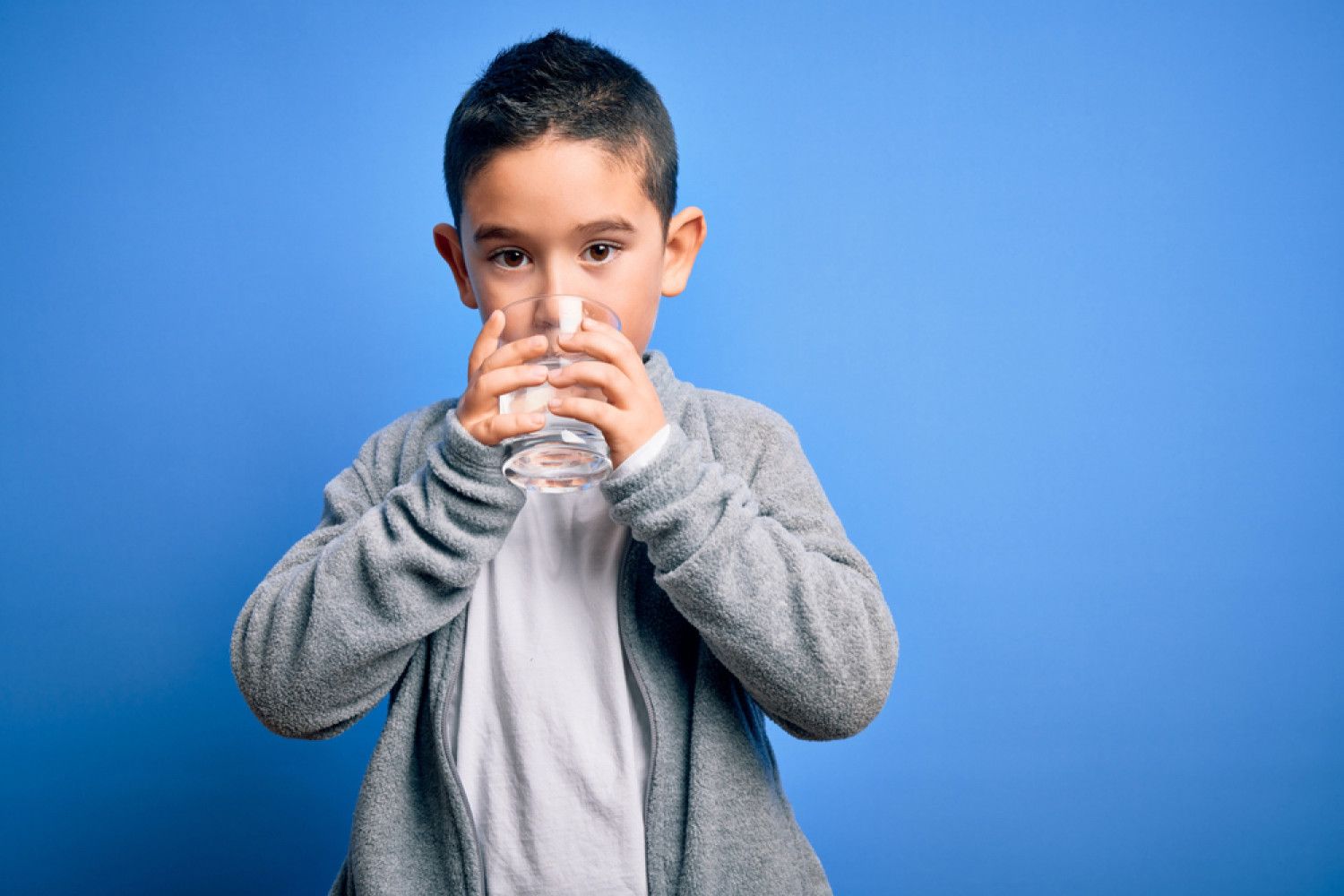 A young boy is drinking a glass of water on a blue background.