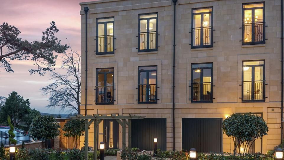 Stone townhouse at dusk with lit windows, framed by trees and a pink-blue sky