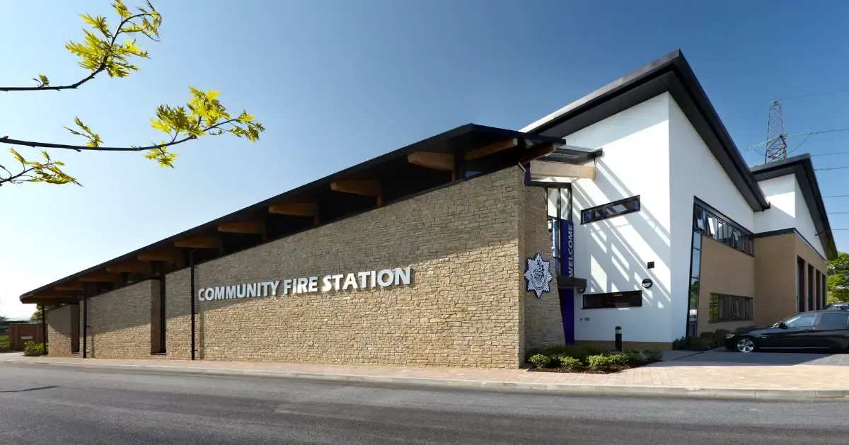 Front view of a modern stone and white fire station building with a sloped roof and parking area.