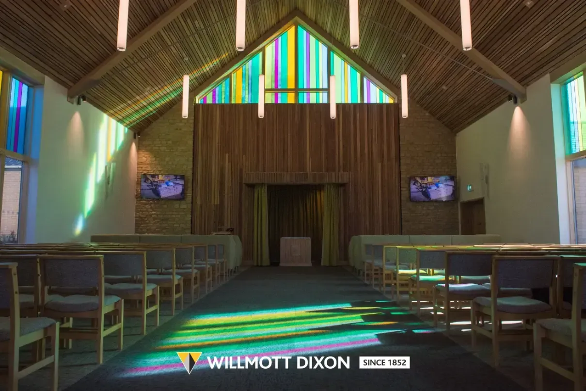 Wooden church interior with rows of pews, colorful stained-glass window, and central aisle leading to the altar