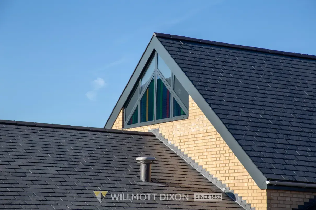 Exterior of a brick building with a dark shingled roof and triangular stained-glass windows against a blue sky