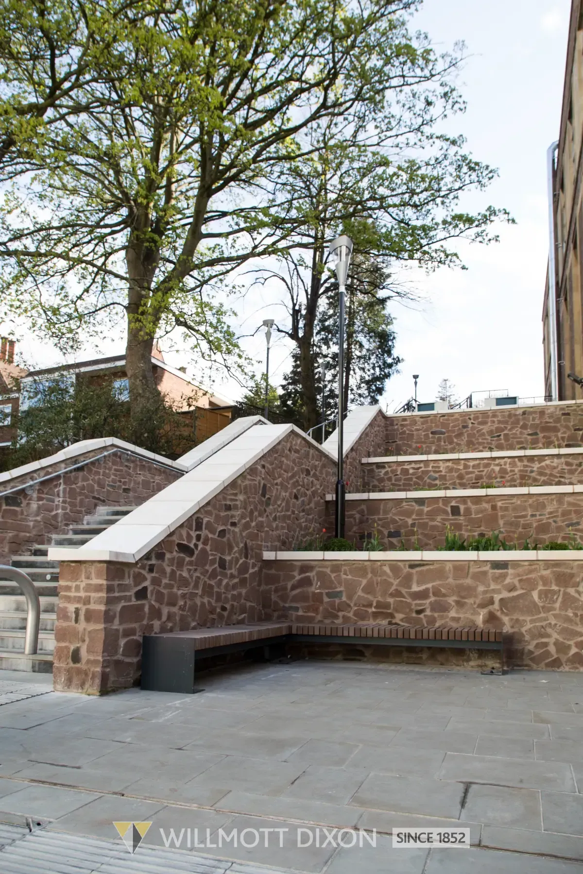 Brick outdoor steps and ramps in a courtyard with trees and a stone bench.