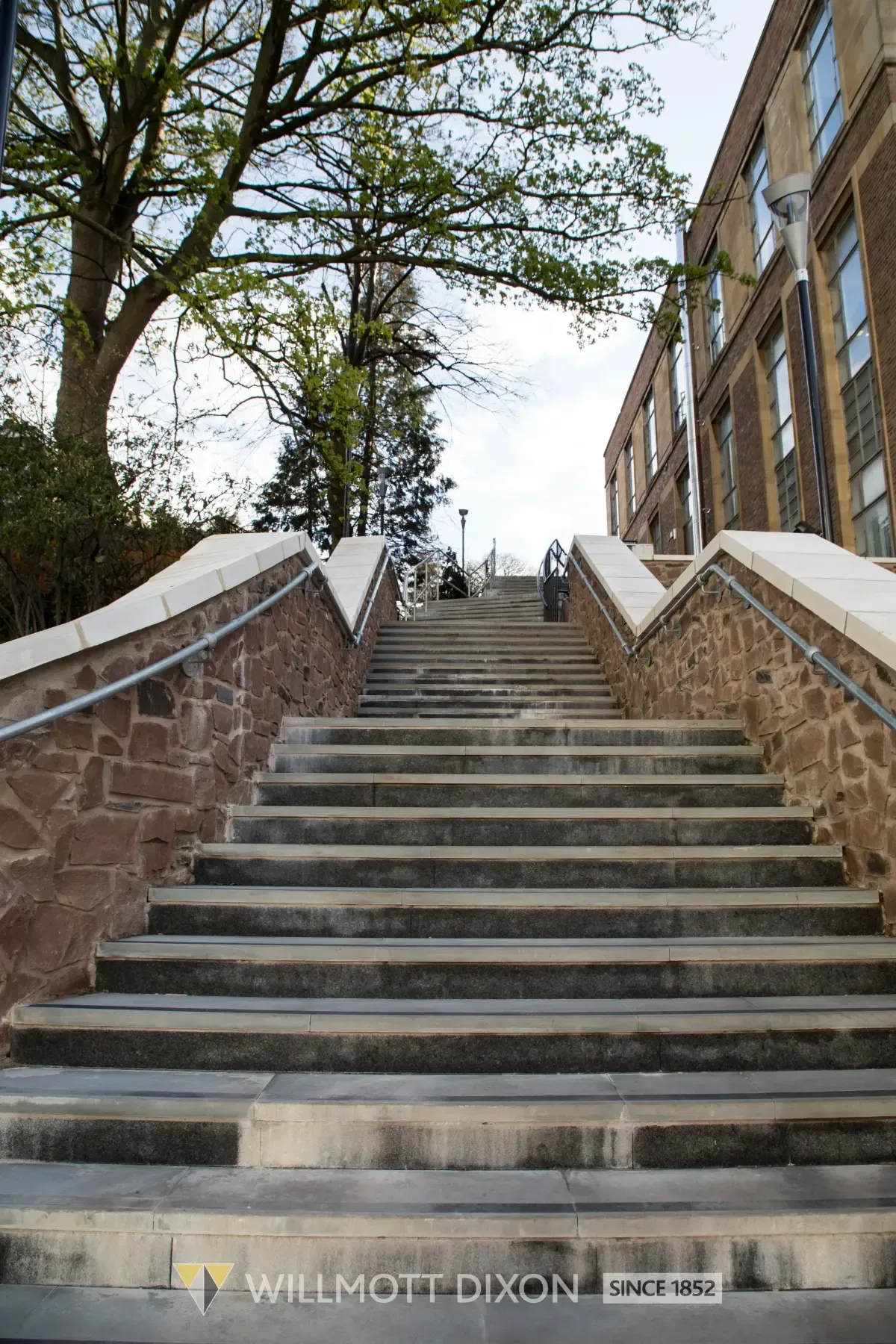 Stone stairway between brick walls leading up to a tree-lined campus path, with a building on the right.