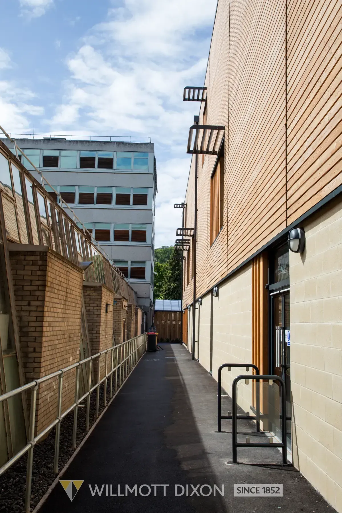 Narrow alley between brick buildings with railings and windows under a partly cloudy sky