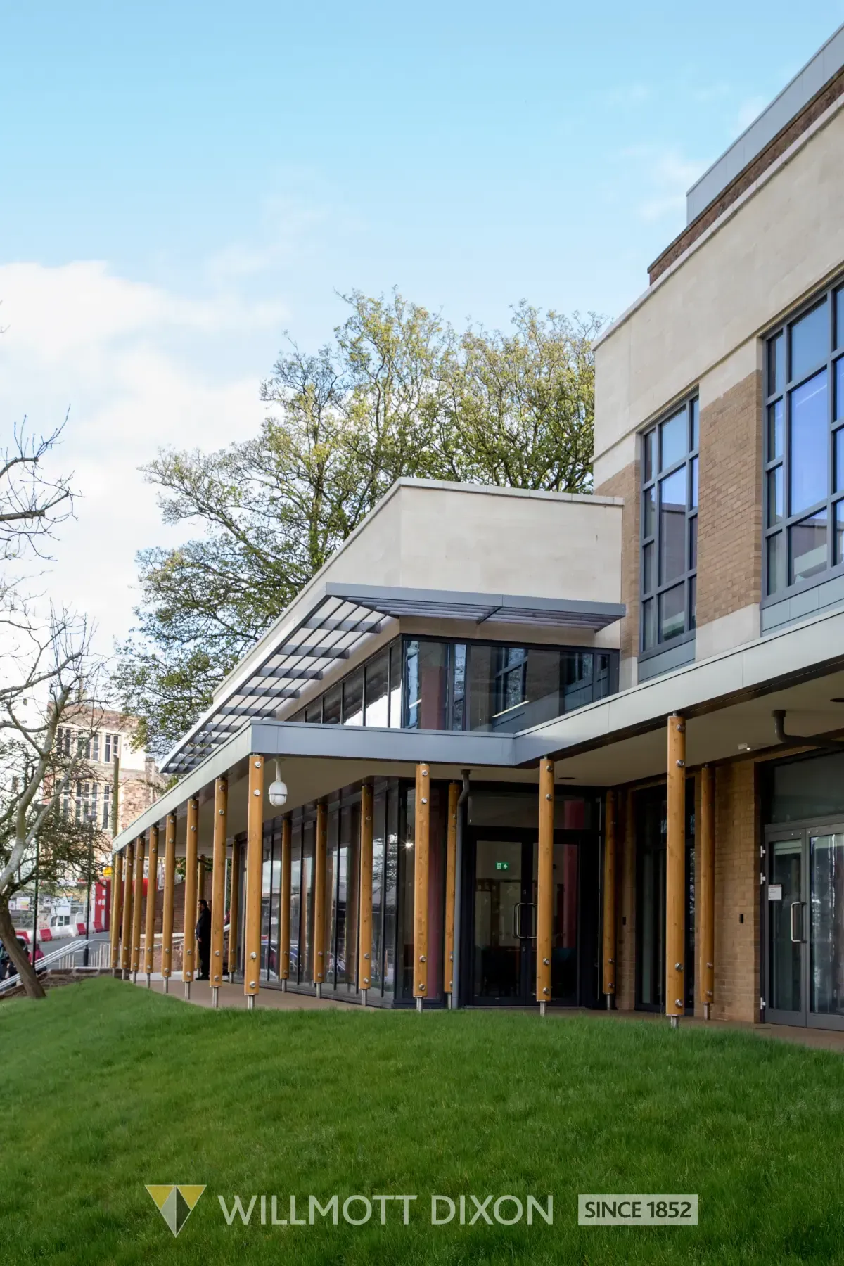 Modern building exterior with glass awning, brick columns, and landscaped walkway under a blue sky