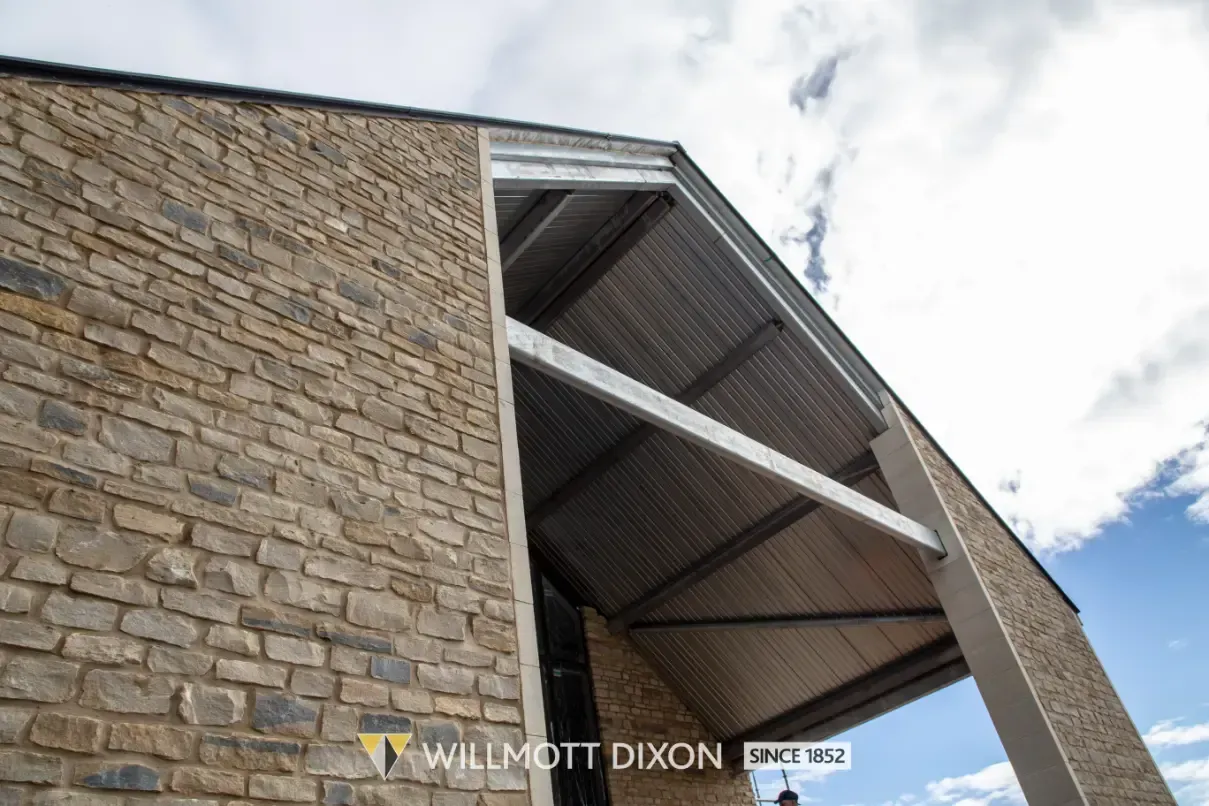 Stone building exterior with a modern covered entrance and slanted metal roof against a cloudy sky