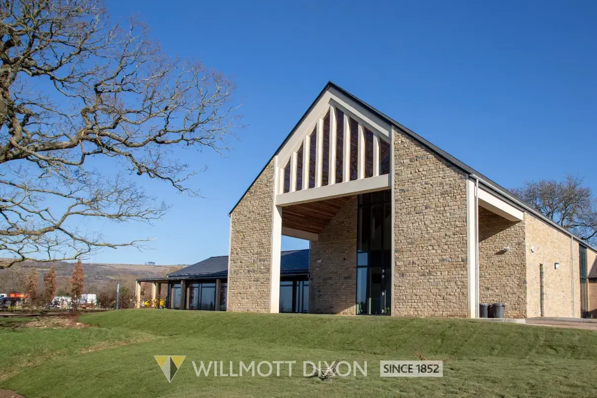 Modern stone building with gabled entrance, large open archway, and grass under a clear blue sky