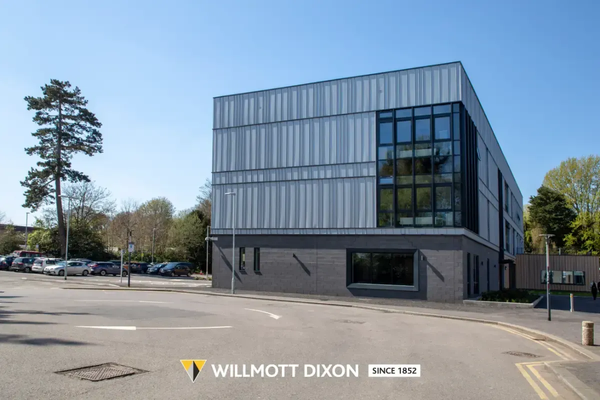 Modern glass-fronted office building on a quiet street with parked cars and trees.