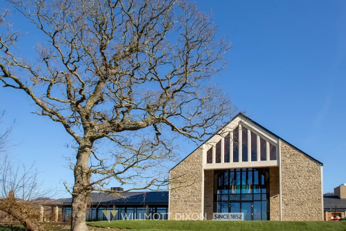 Stone building with a peaked roof and large windows beside a leafless tree under a clear blue sky