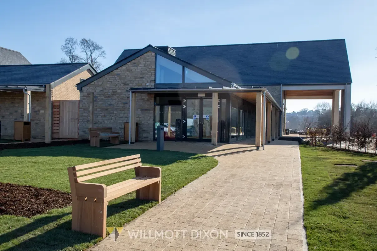 Modern brick building with glass entrance, paved path, and a wooden bench on a lawn