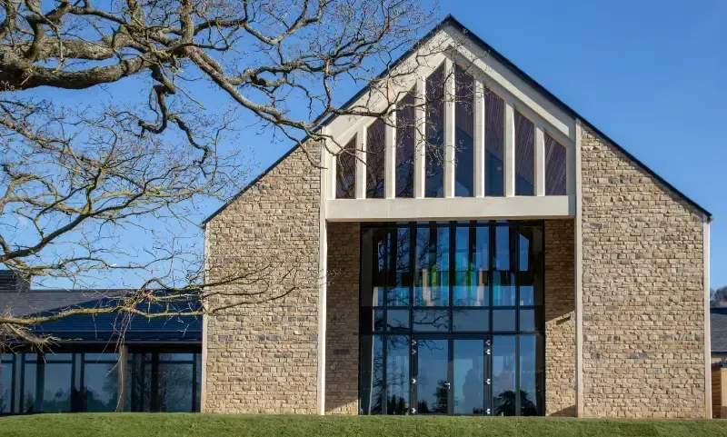 Stone building with tall glass entrance and triangular roof, framed by bare tree branches under a blue sky