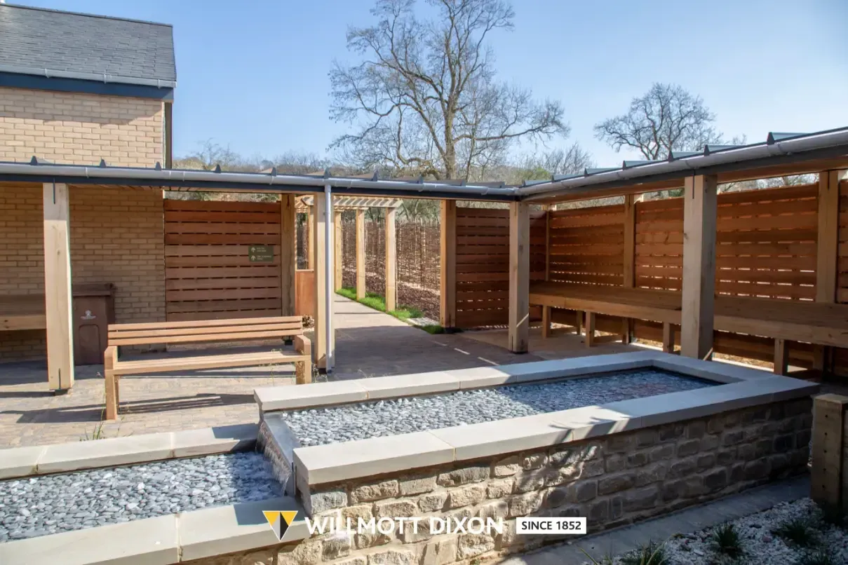 Outdoor courtyard with wooden benches, covered seating areas, and a brick planter in sunlight