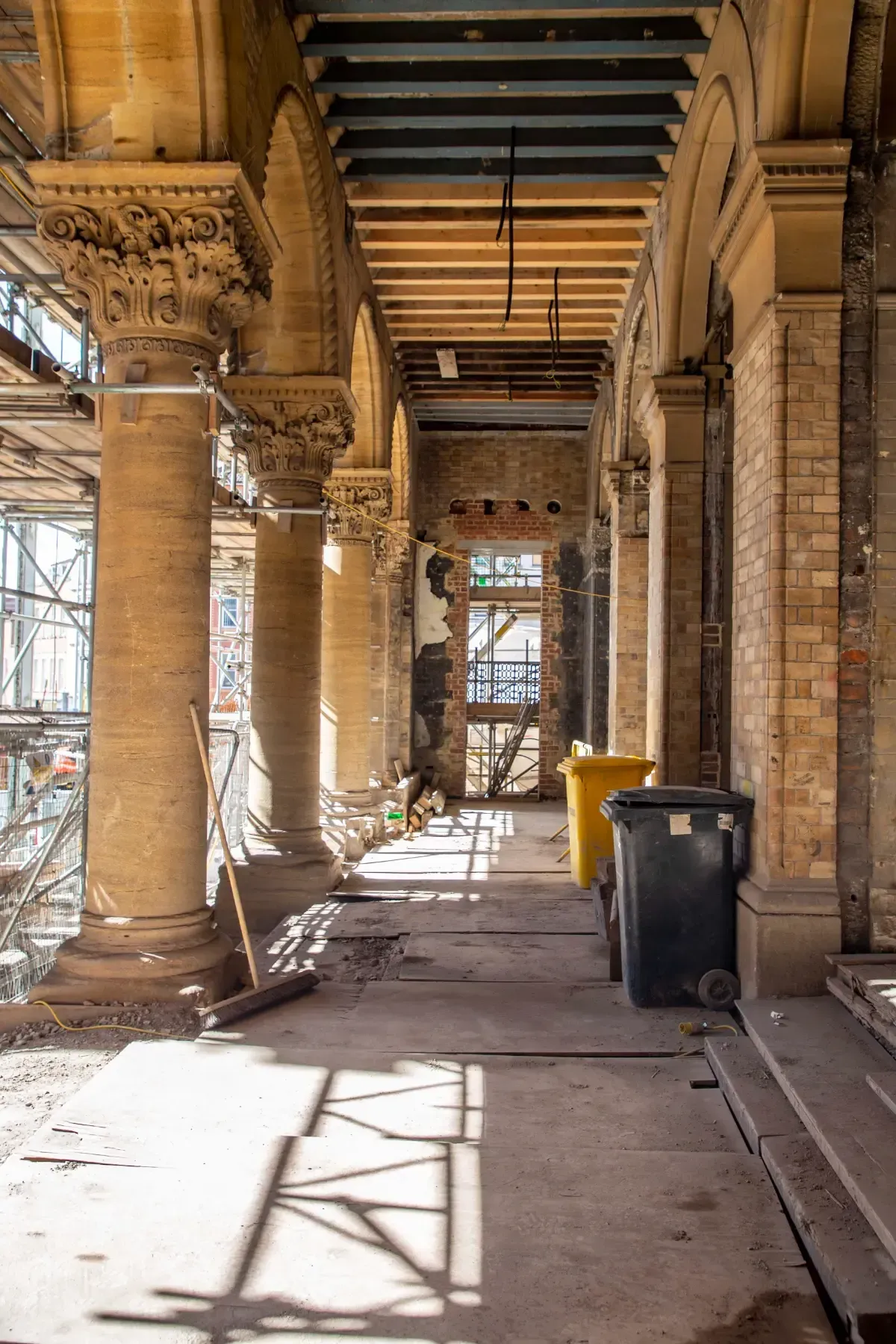 Stone colonnade under renovation with scaffolding, sunlight, and a trash bin along a covered walkway