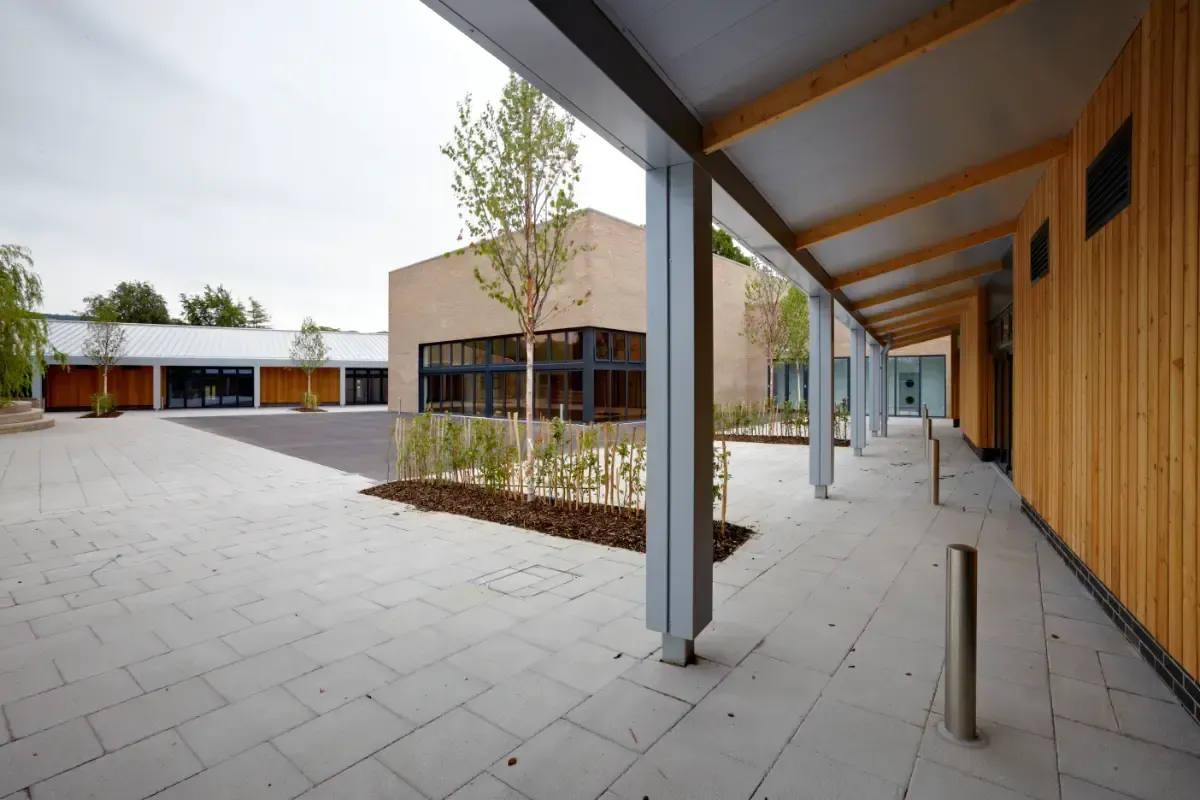 Covered walkway in a modern courtyard with brick buildings, trees, and a paved open plaza