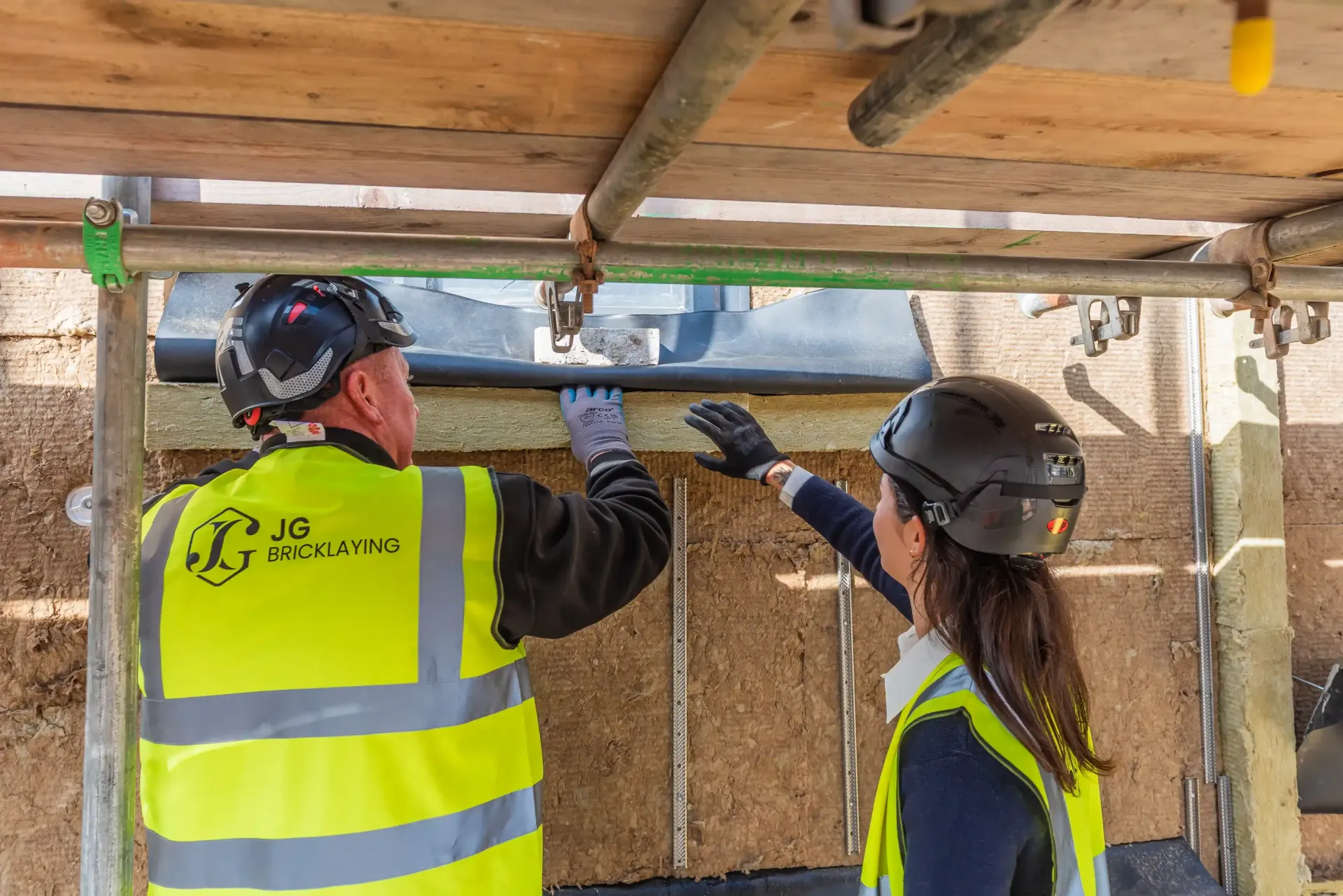 Two construction workers in hard hats and hi-vis vests inspect a wall under a scaffold.
