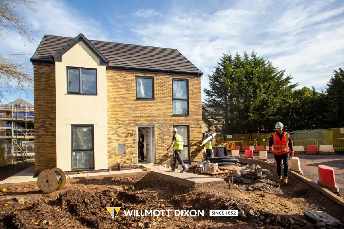 Two workers in orange vests at a muddy house construction site with a yellow-brick home.