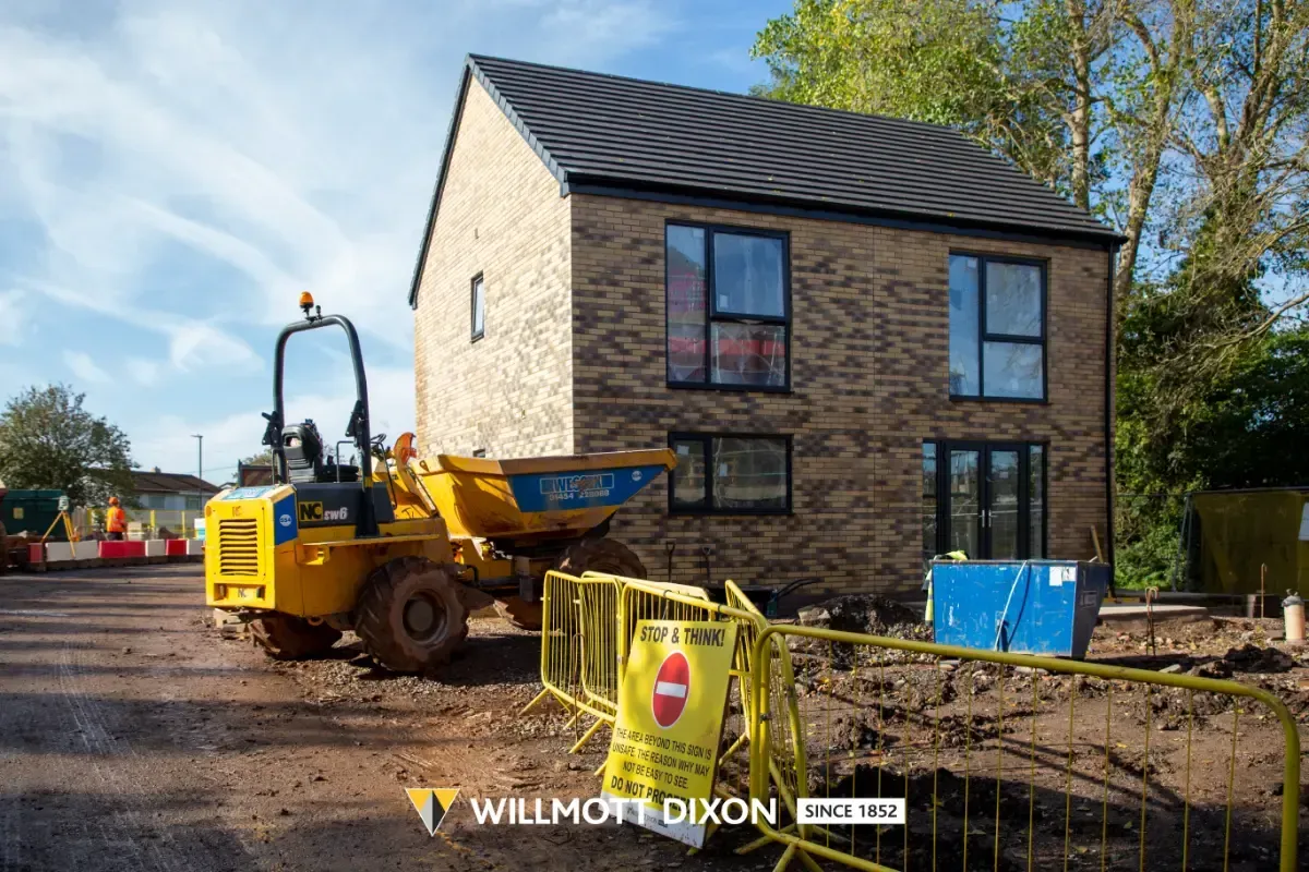 Yellow construction vehicle outside a stone house under construction, with muddy ground and fencing in front