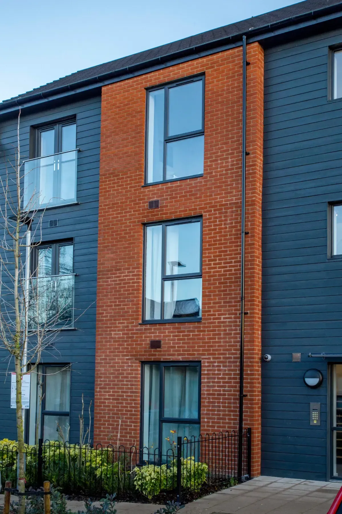Modern apartment building exterior with red brick and dark blue siding, black railings, and balconies.