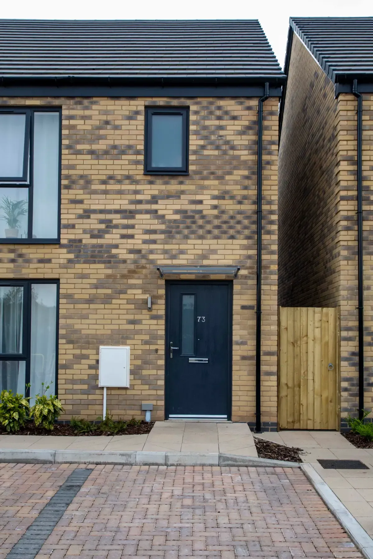 Modern brick townhouse entrance with dark blue door, narrow window, and wooden side gate