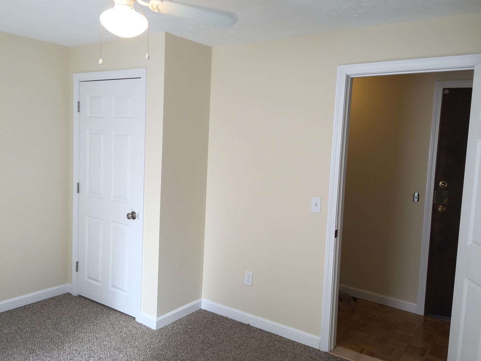 Empty room with closed white door, open doorway, tan walls, and carpet.