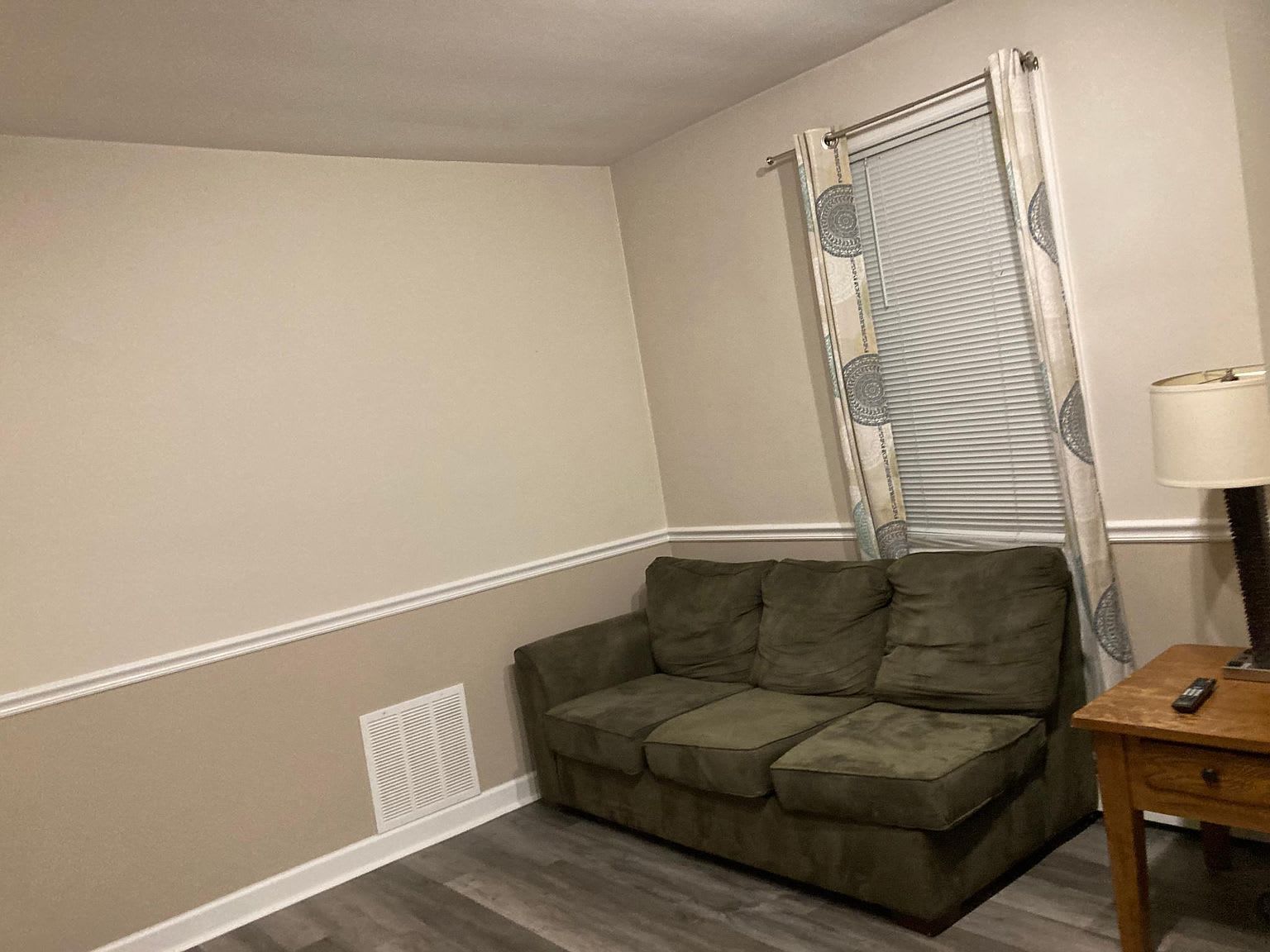 Green couch in a room with a window, curtains, and a side table with a lamp.