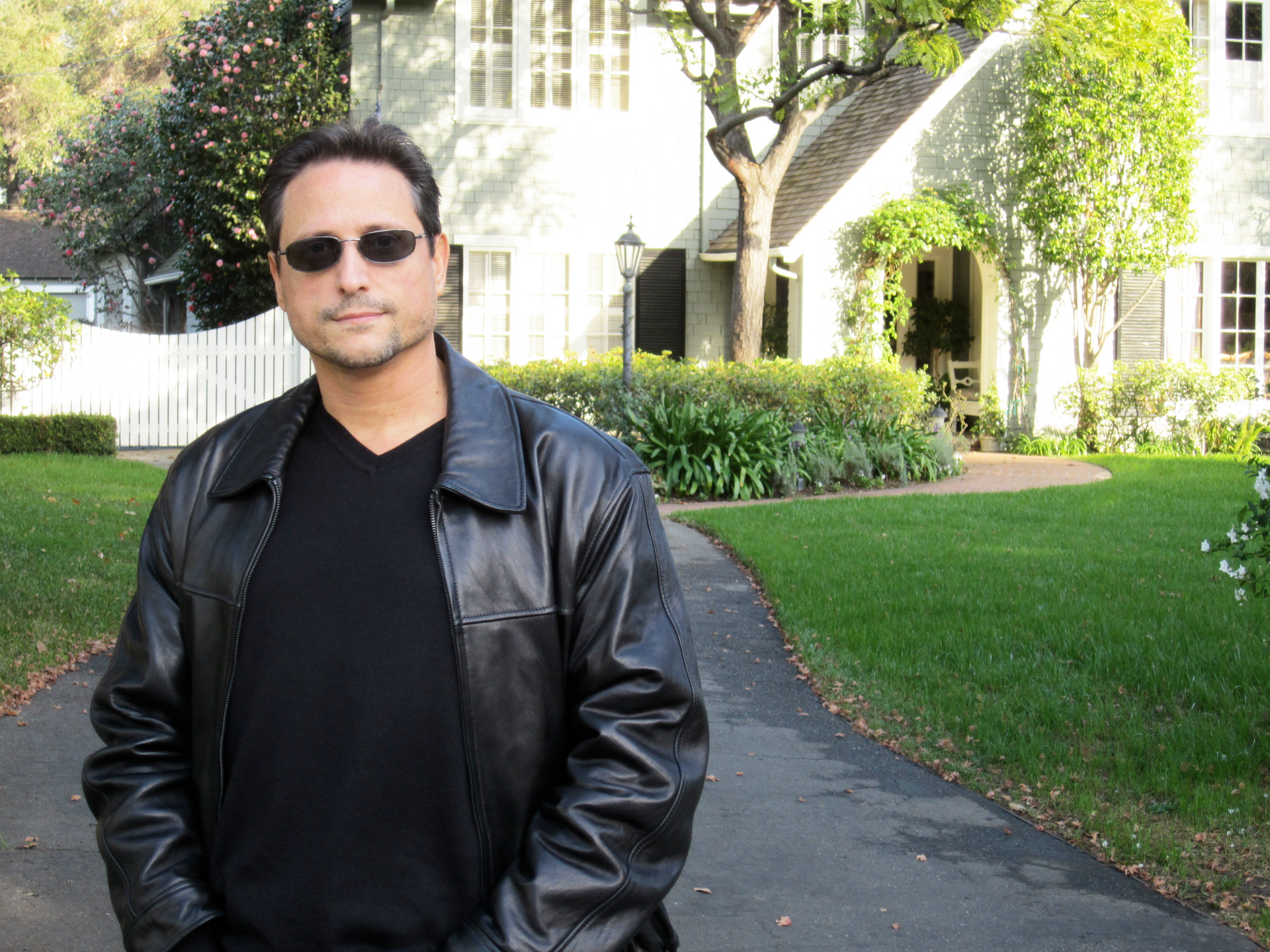 Al Conti in black leather jacket and sunglasses in front of house.