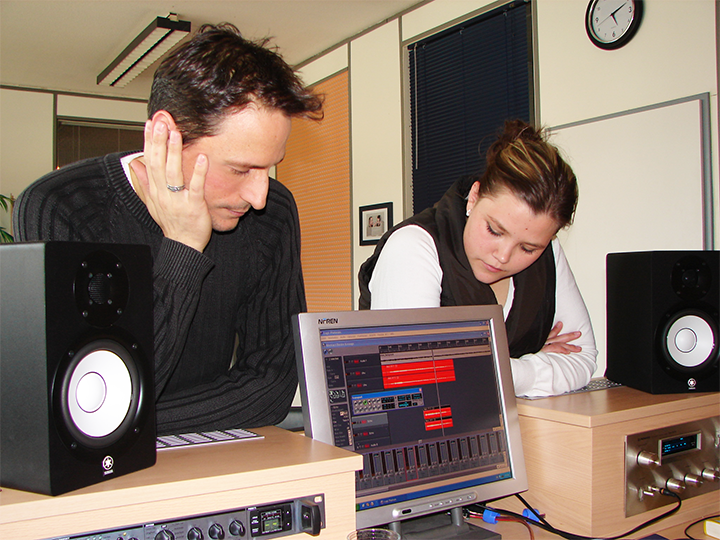 Two people in a recording studio, looking at a computer screen with audio software. Black speakers sit on either side.
