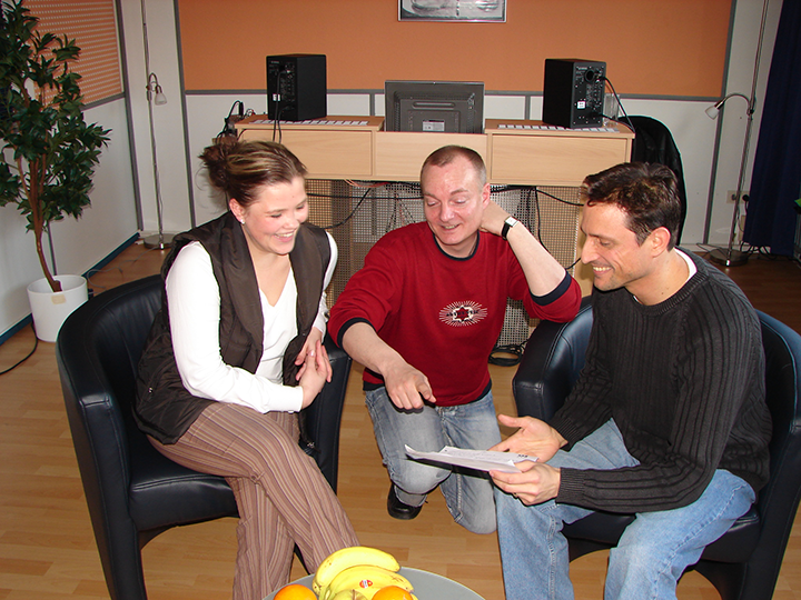 Three people in a studio discussing paperwork; woman smiles, man in red gestures, man in black holds document.