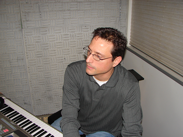 Al sits near a keyboard, looking to the side. Gray shirt, textured wall background.