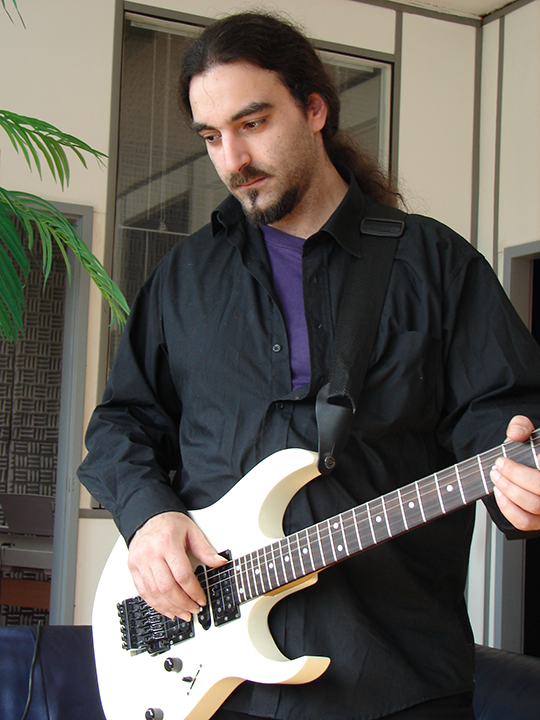 Man with long hair plays a white electric guitar, wearing a black shirt. Indoors.