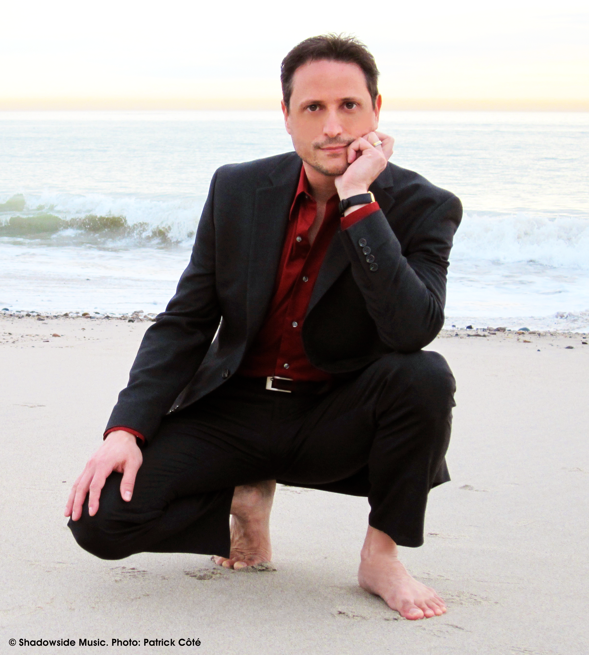 Al Conti barefoot on beach, resting chin on hand. Red shirt, dark jacket.