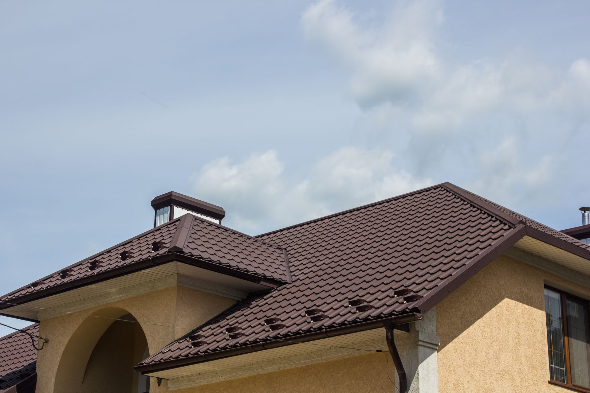 Dark brown metal tile roof on stucco house with chimney against blue sky.
