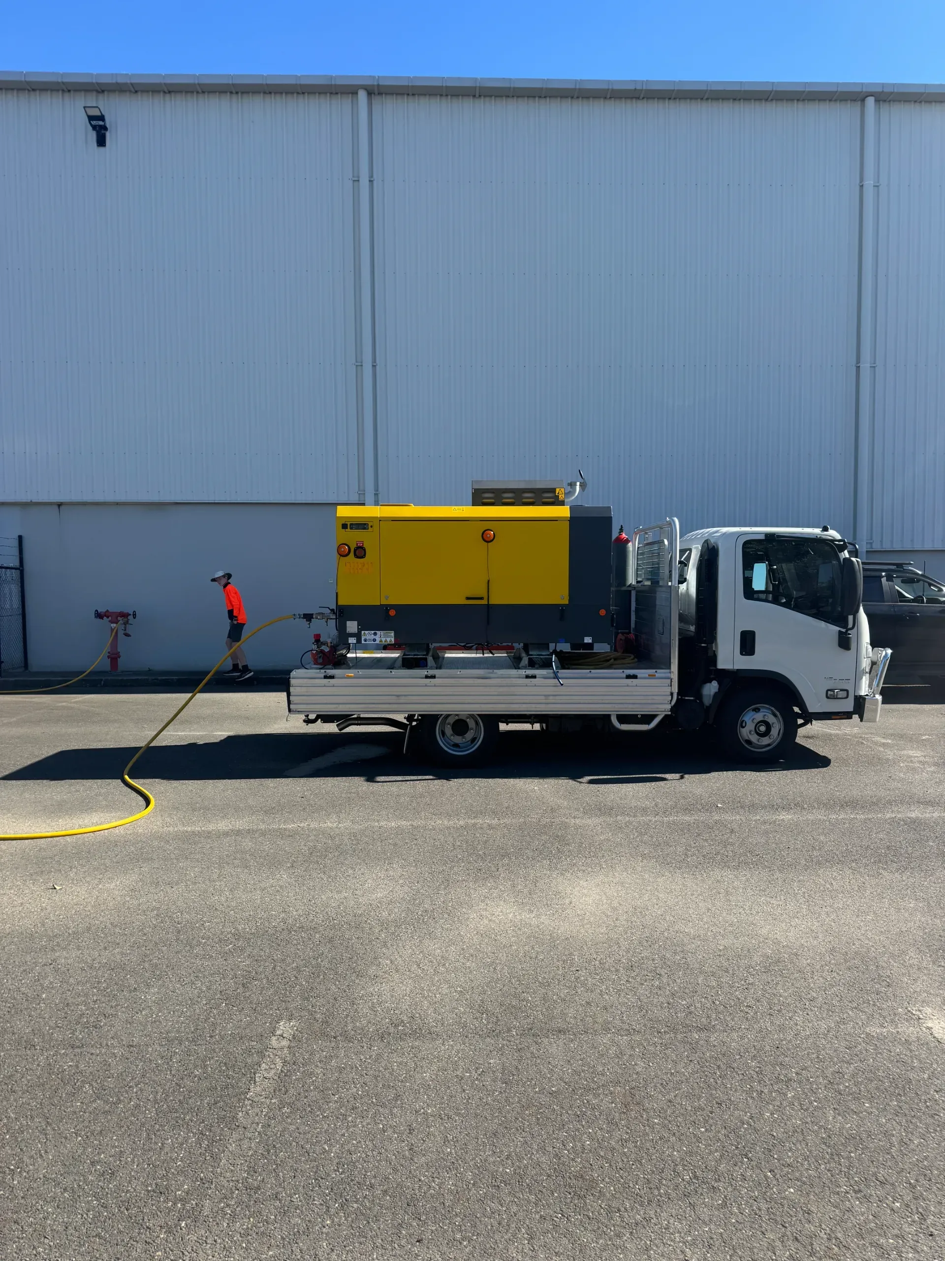 Truck with yellow equipment, worker holding hose, parked in front of a white building on a sunny day. — Commercial Leak Detection