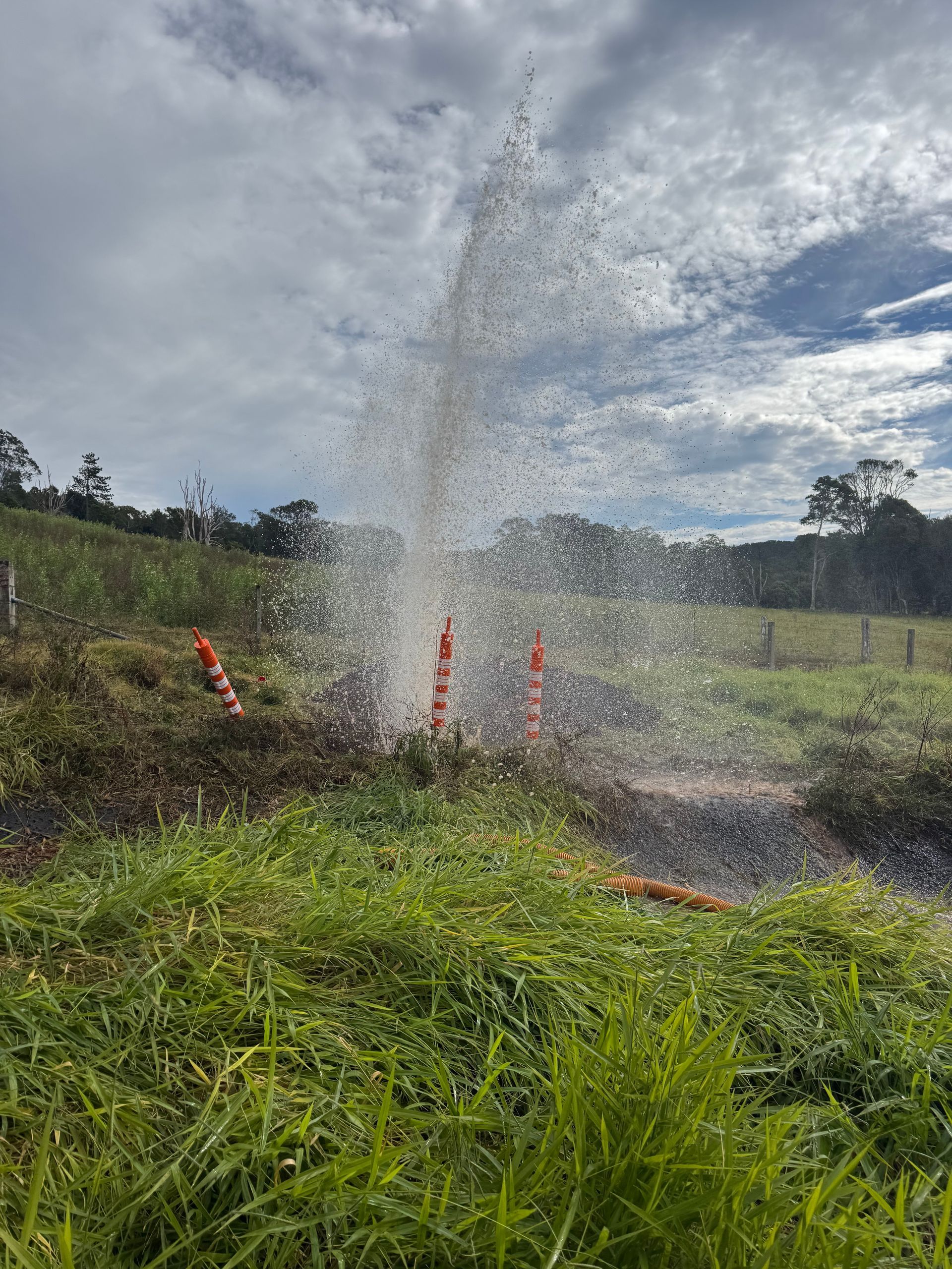 Water erupts from a pipe in a grassy field, surrounded by orange cones under a cloudy sky.  Commercial Leak Detection in Tweed Heads, NSW