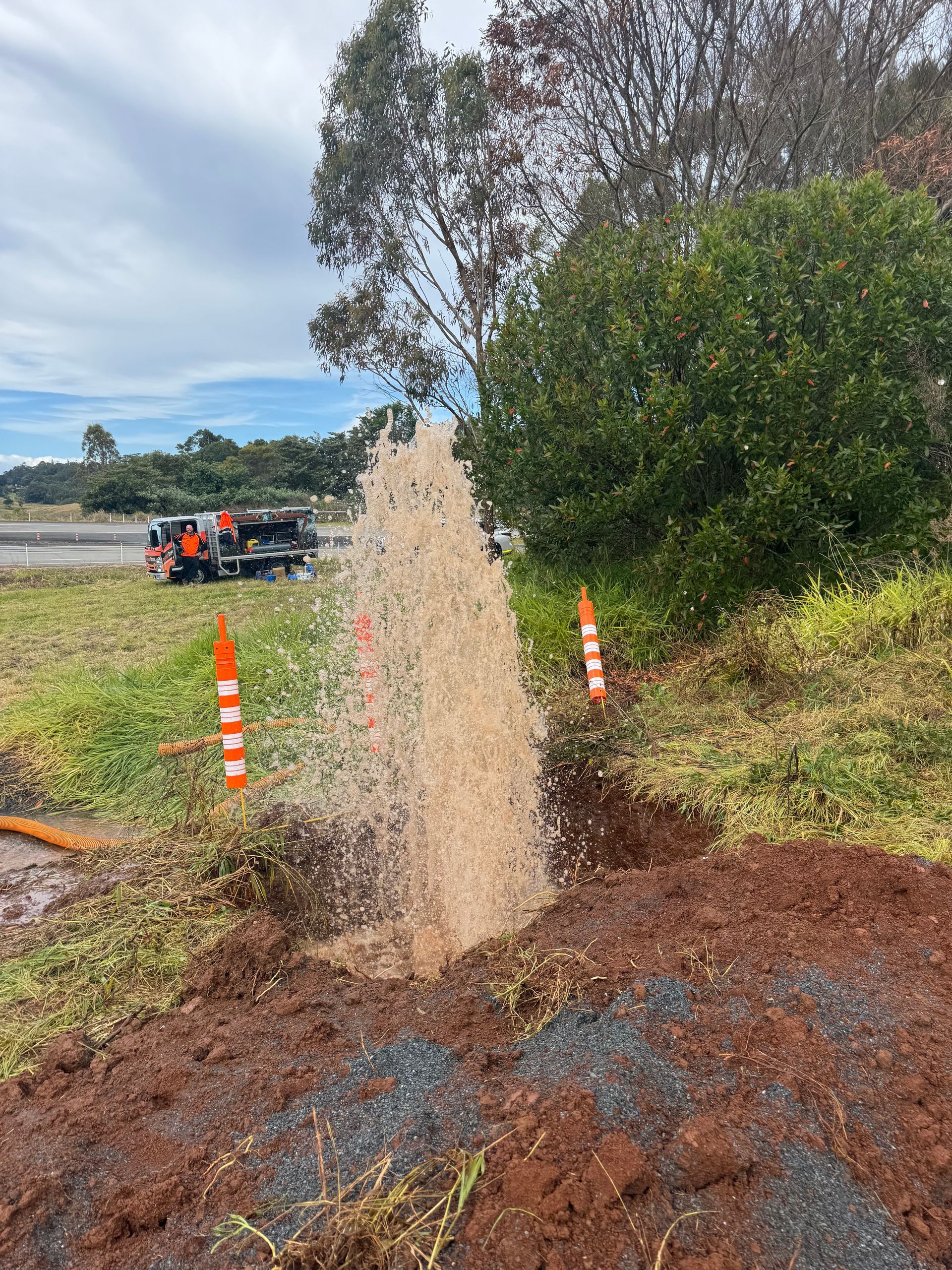 Water gushing from a break in a roadside pipe. Brown water sprays upwards. Orange cones and a truck are in the background.