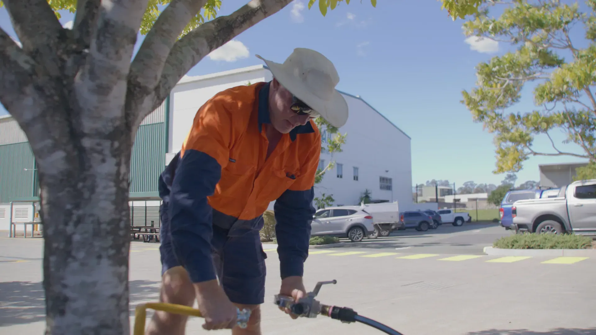 Man in work clothes uses a hose near a tree in a parking lot on a sunny day. — Commercial Leak Detection