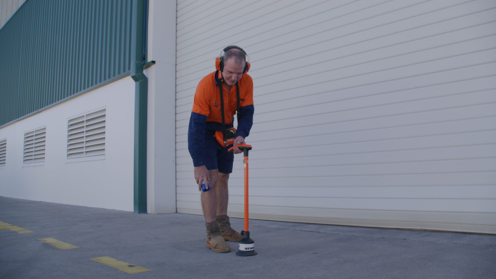 Man in work clothes uses a line marking machine on an asphalt surface. White building in background. — Commercial Leak Detection in Arundel, QLD