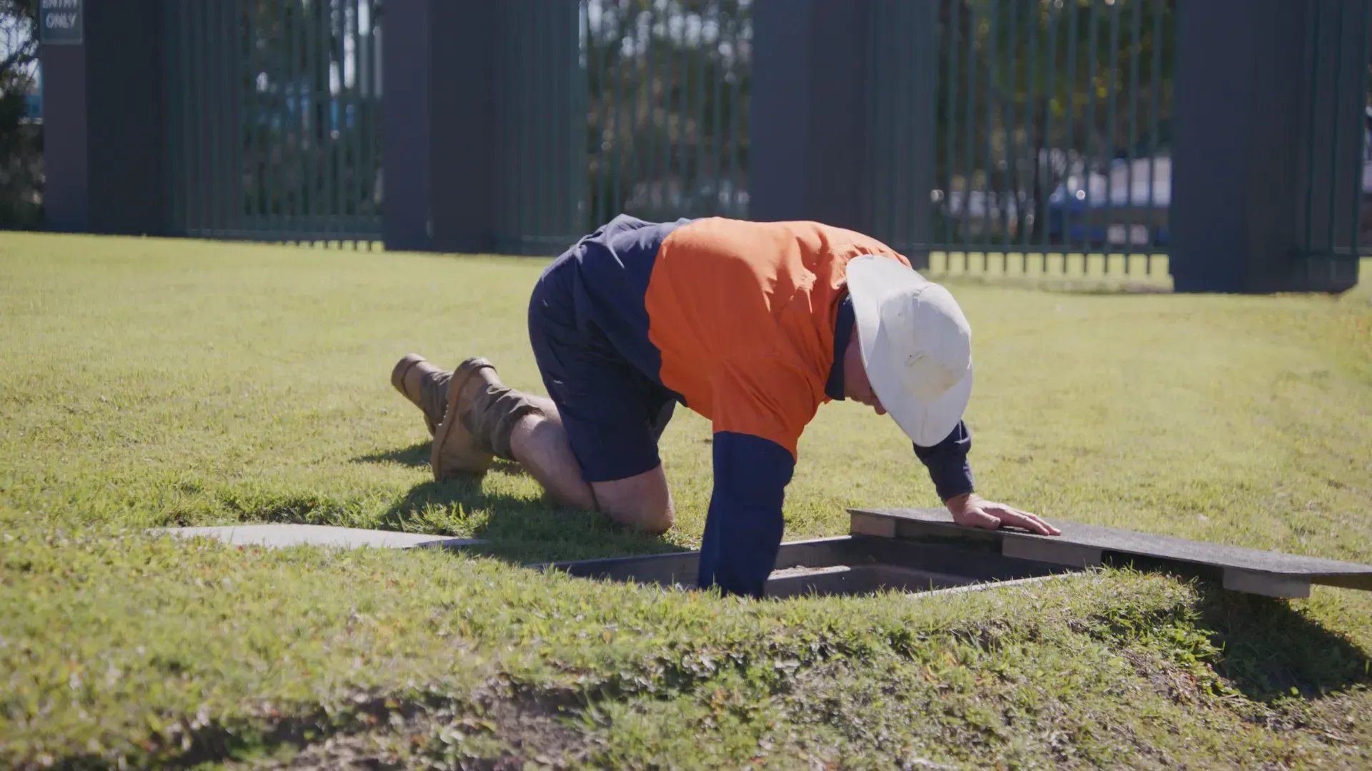 Construction worker kneels, looking into a rectangular hole in the grass; wearing an orange shirt and white hat. — Commercial Leak Detection