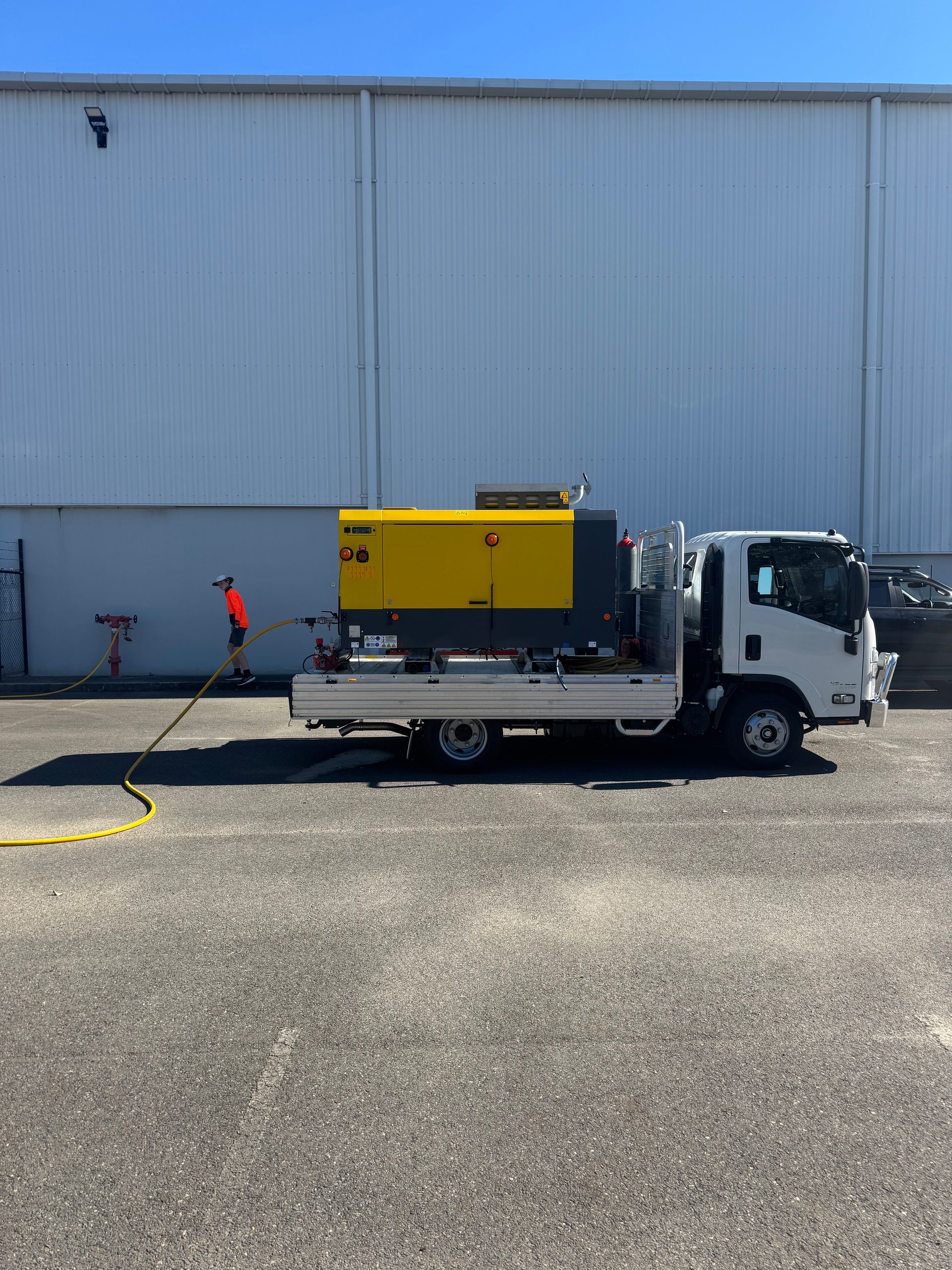 A truck with yellow equipment, a person in orange, and a long yellow hose on asphalt. Building in the background.  — Commercial Leak Detection in Burleigh Heads, QLD
