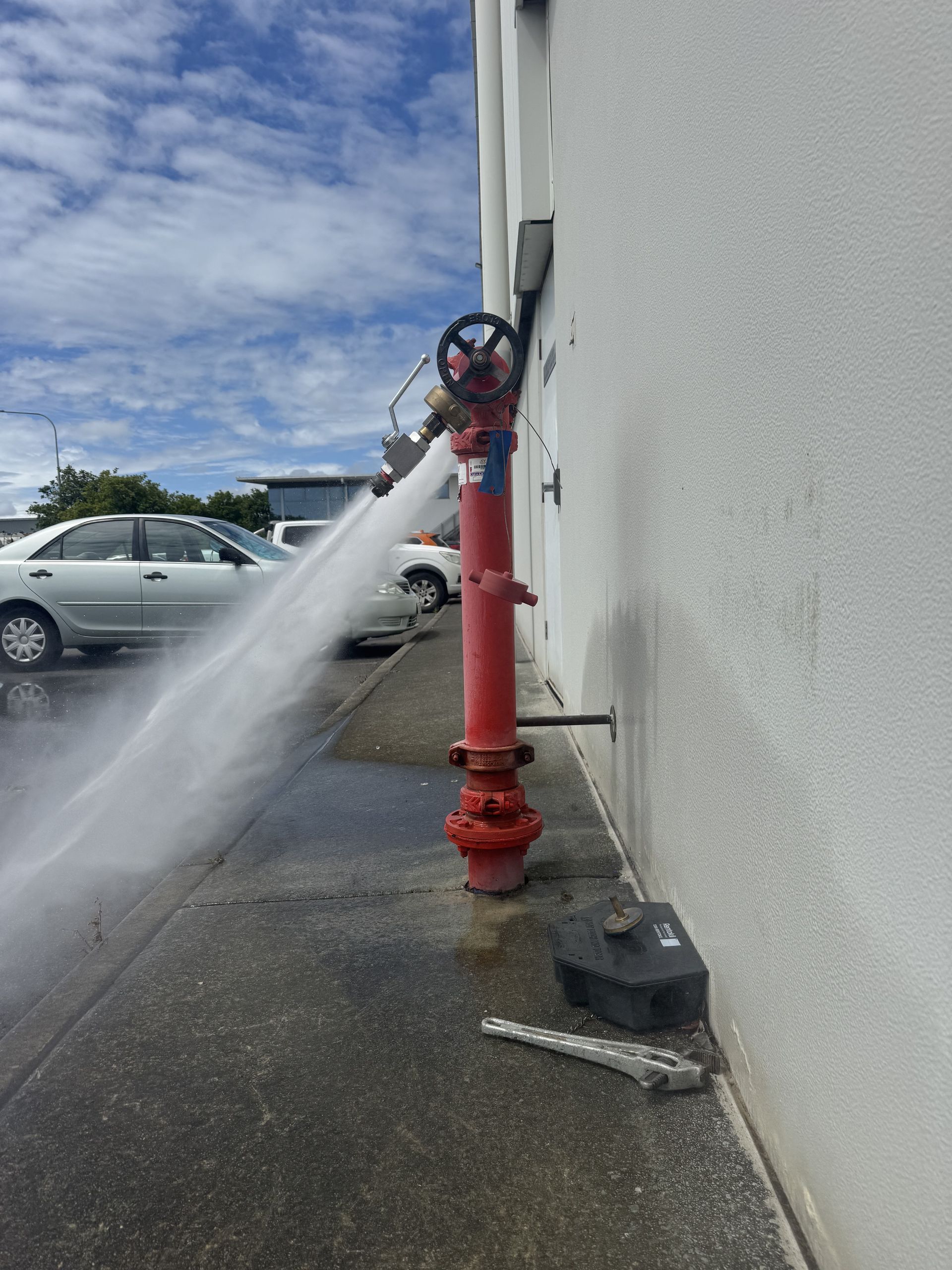 A red fire hydrant spurting water beside a white building and parked cars under a cloudy sky.