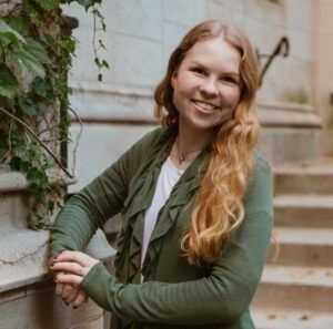 Smiling person with long red hair leaning against a stone wall in a green jacket and white shirt