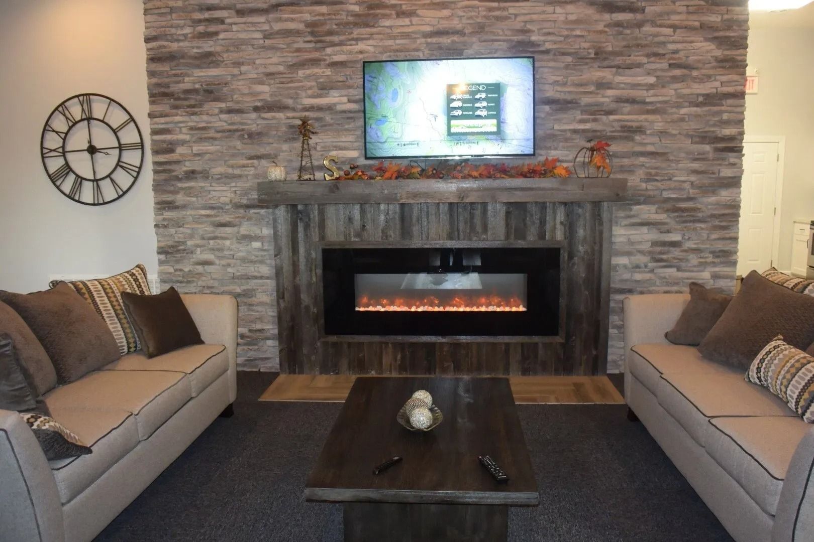Cozy living room with stone fireplace, wall-mounted TV, two sofas, coffee table, and decorative wall clock.